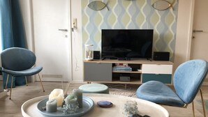 En moderne stue med et fladskærms-tv, to blå lænestole, et sofabord med stearinlys og en bogreol.