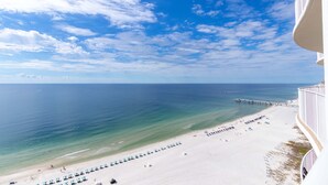 A beach with a pier extending into the ocean, a row of beach umbrellas, and a white building with balconies overlooking the beach.