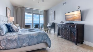 A bedroom with a bed, a desk, a dresser, and a large window with a view.