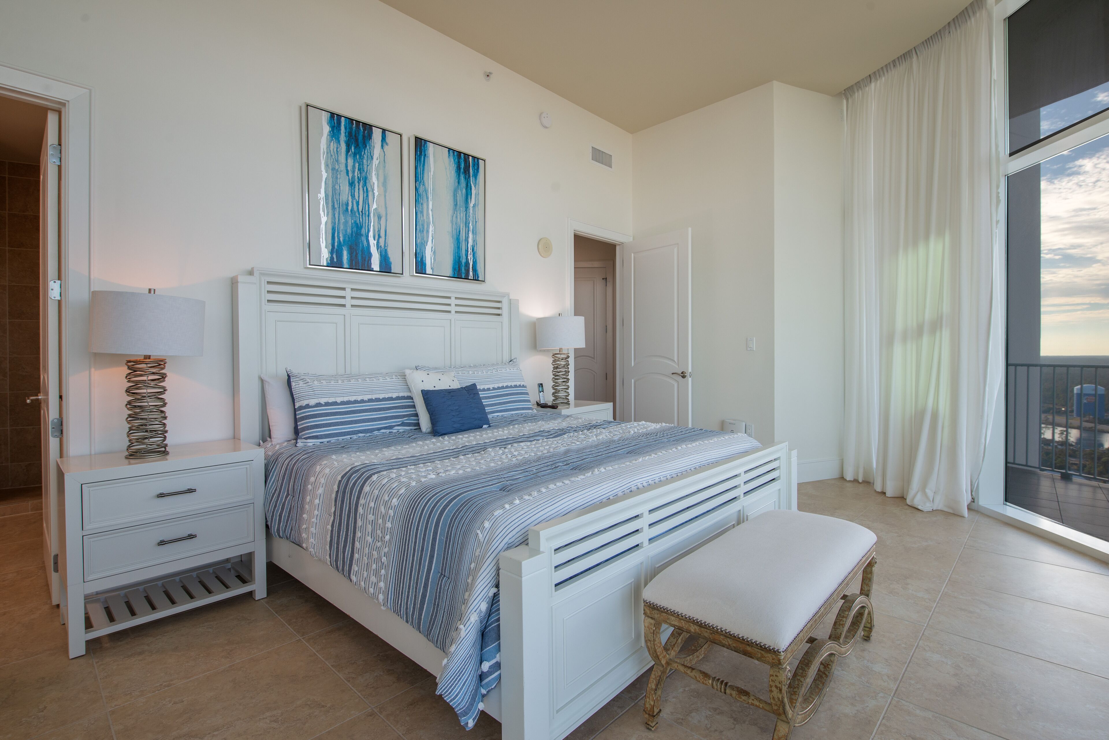 Un dormitorio con cama, mesitas de noche, un banco y un amplio ventanal con cortinas.