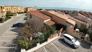 Un quartier résidentiel avec une voiture blanche garée dans une allée, un bâtiment avec un toit en tuiles, et une vue sur la mer en arrière-plan.