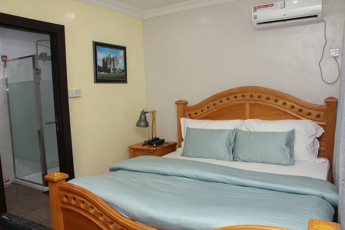 A bedroom with a wooden bed, a bedside table with a lamp, a framed picture on the wall, and an air conditioning unit.