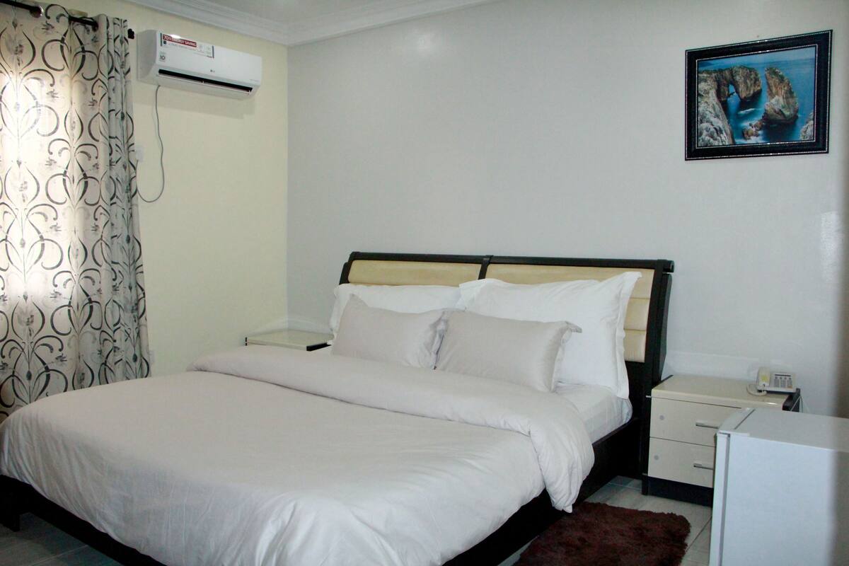 A neatly made bed with white linens, a bedside table with a lamp, and a wall-mounted air conditioner.