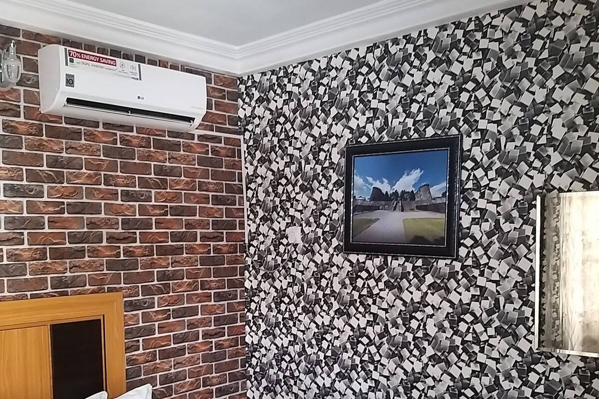 A hotel room with a brick-patterned wall, a bed, a desk, and an air conditioning unit.