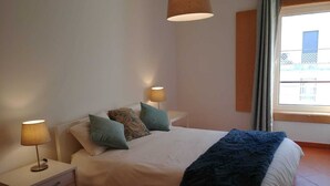 A bedroom with a bed, two bedside tables with lamps, a window with curtains, and a view of buildings outside.