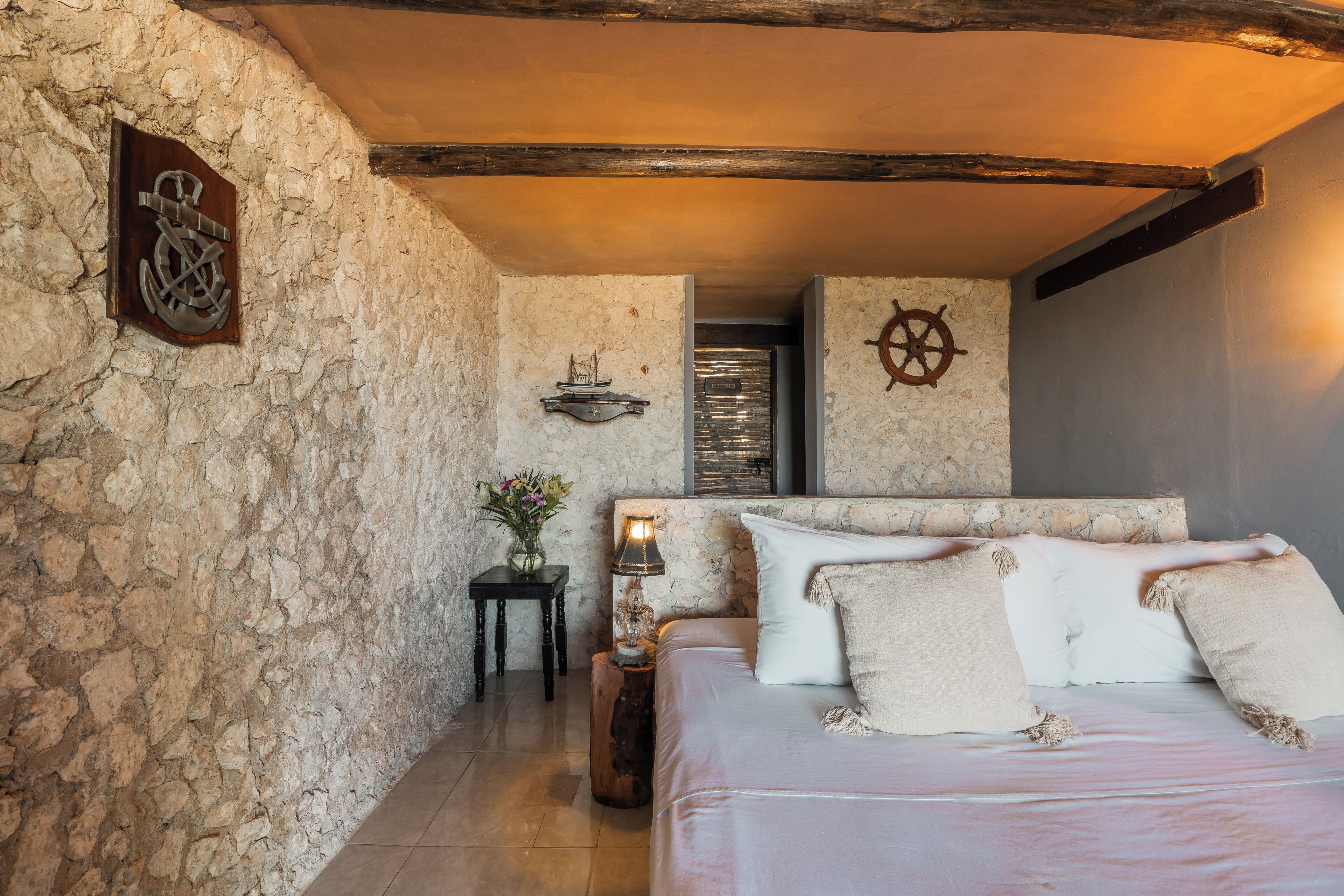 Une chambre avec un mur en pierre, un plafond en bois, un lit recouvert de draps blancs et une petite table avec une lampe.