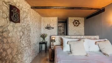 Une chambre avec un mur en pierre, un plafond en bois, un lit recouvert de draps blancs et une petite table avec une lampe.