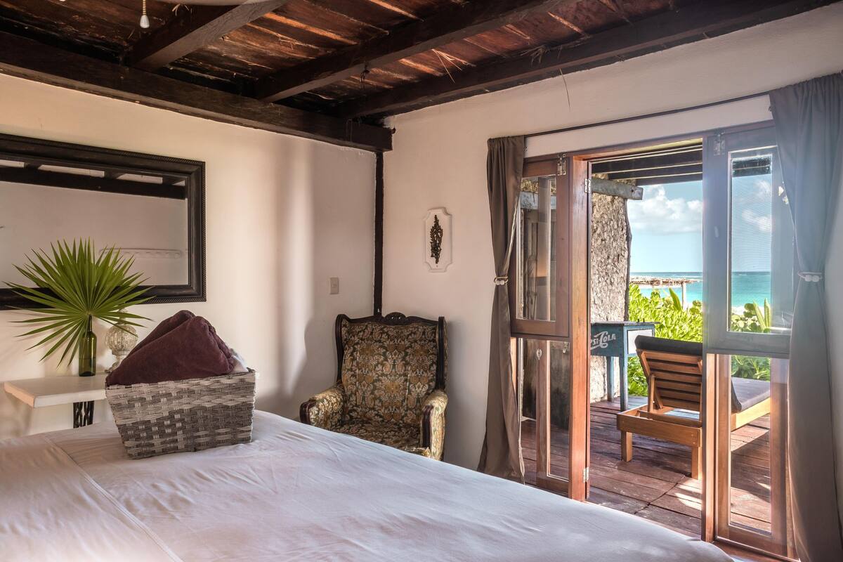A bedroom with a bed, a chair, a plant, and a view of the ocean through a sliding door.