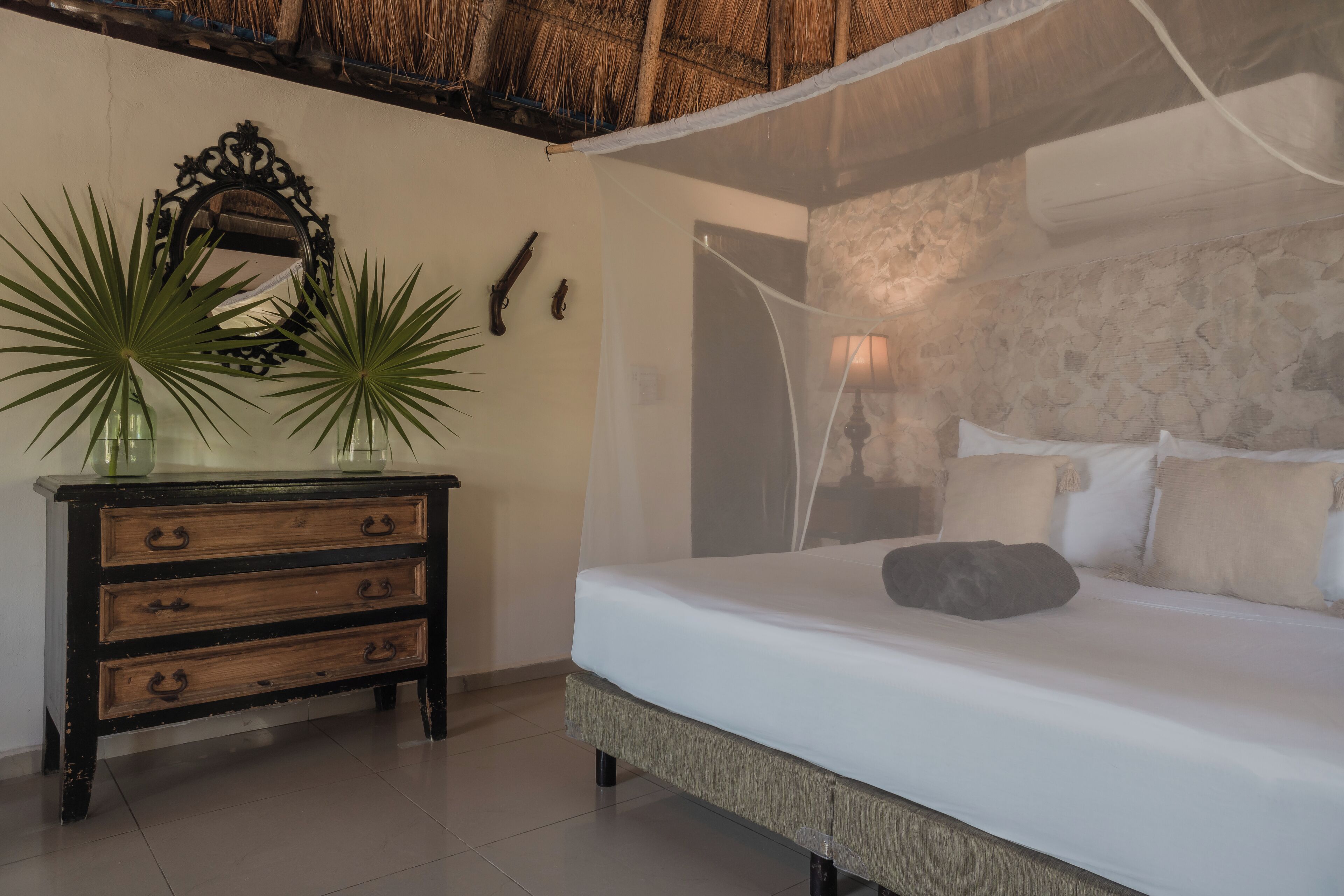 Un dormitorio con una cama, un aparador de madera con cajones, un espejo y dos plantas en macetas.