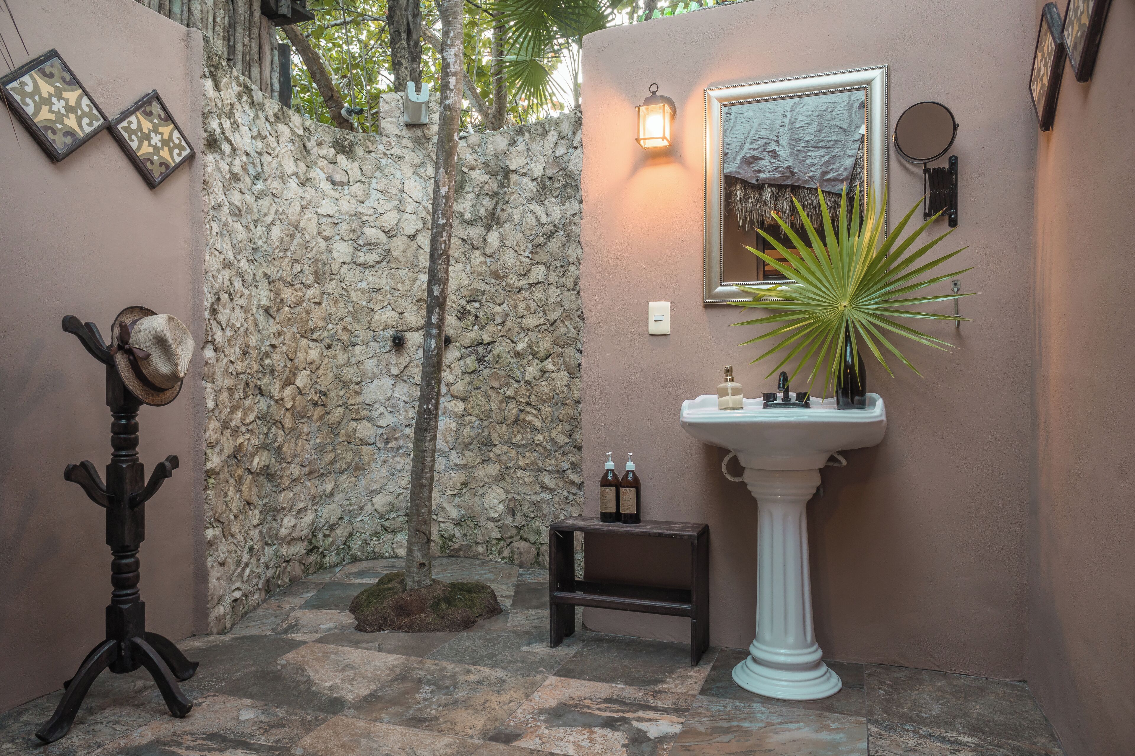 Un baño con pared de piedra, un lavamanos blanco pedestal, un espejo, un estante pequeño con botellas y un sombrero decorativo sobre un soporte.