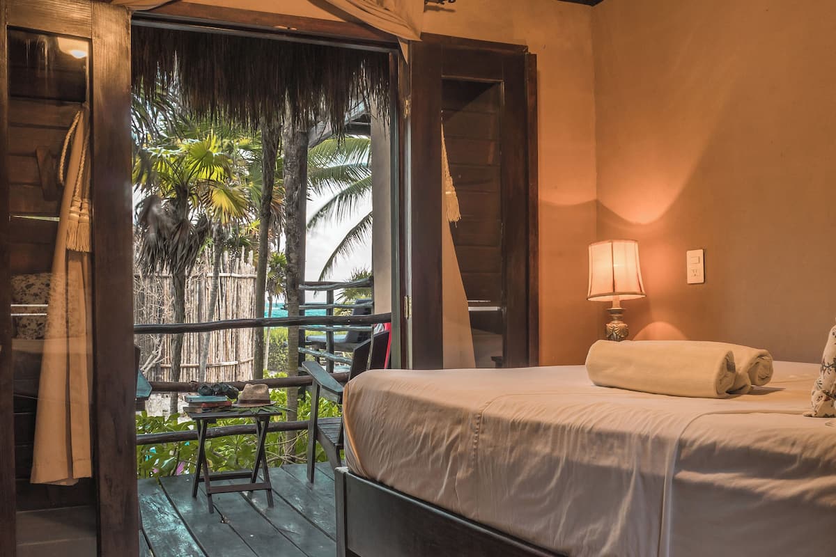 A room with a bed, a nightstand, a lamp, and a view of a palm tree through an open door.
