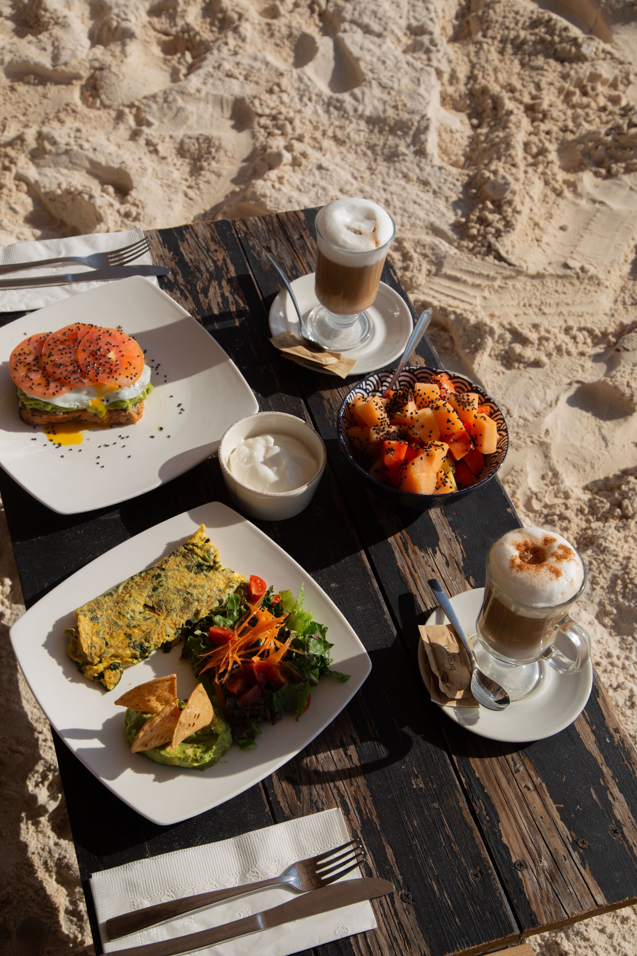 A table with food and drinks on a wooden surface, with a sandy background.
