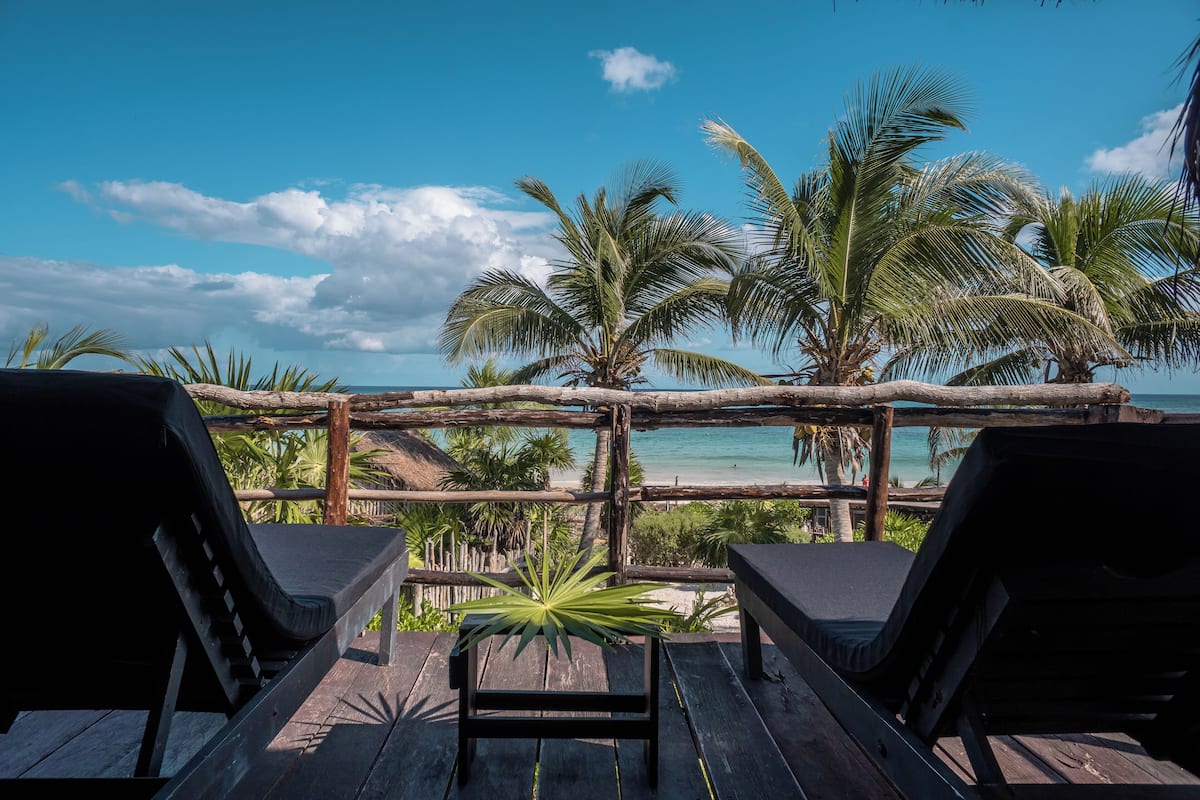 A wooden deck with two lounge chairs facing a beach with palm trees and a wooden fence.