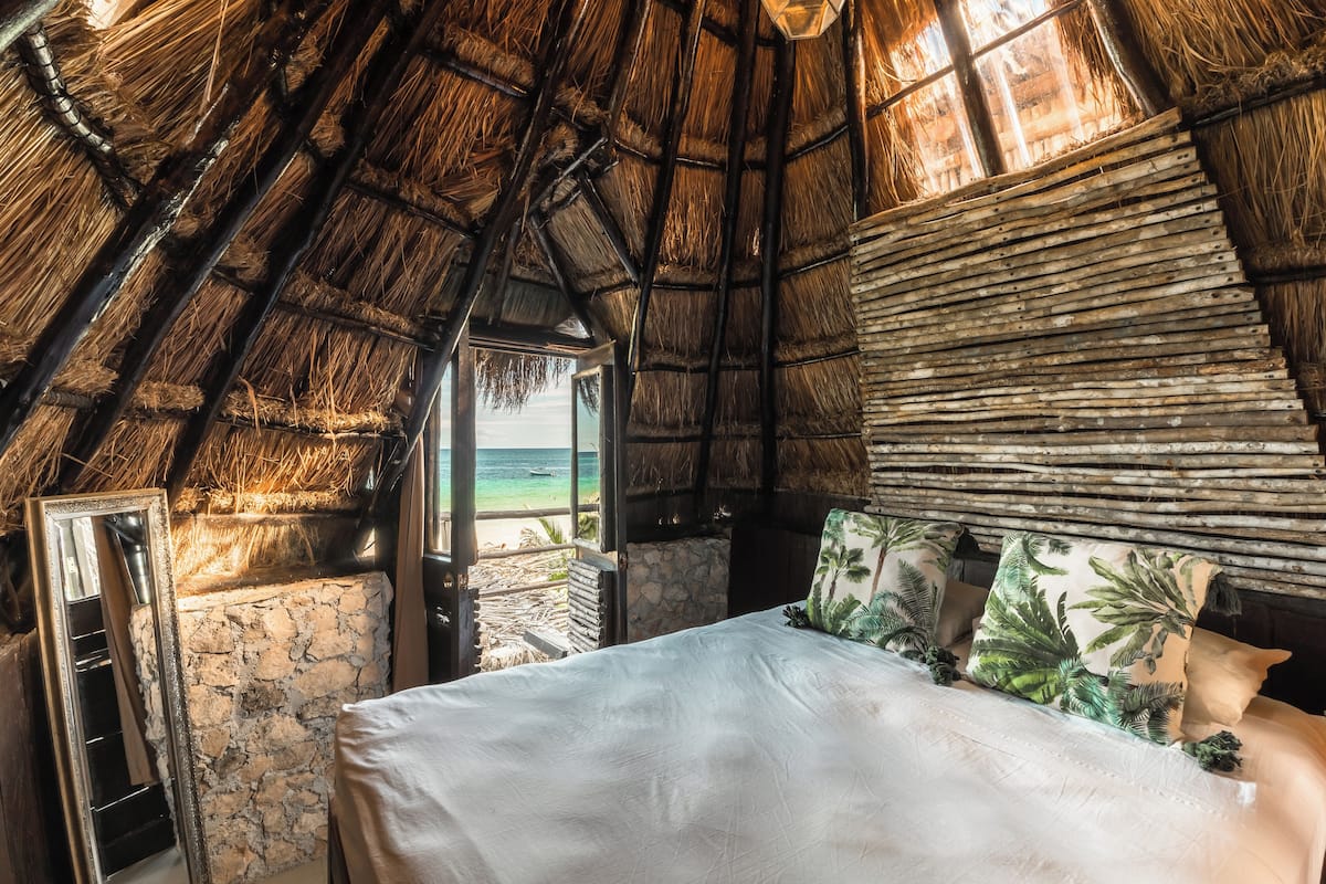 A room with a thatched roof, a bed with white linens, and a view of the beach.