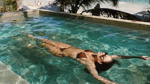Una piscina cristalina con una persona nadando, rodeada de palmeras y una playa al fondo.