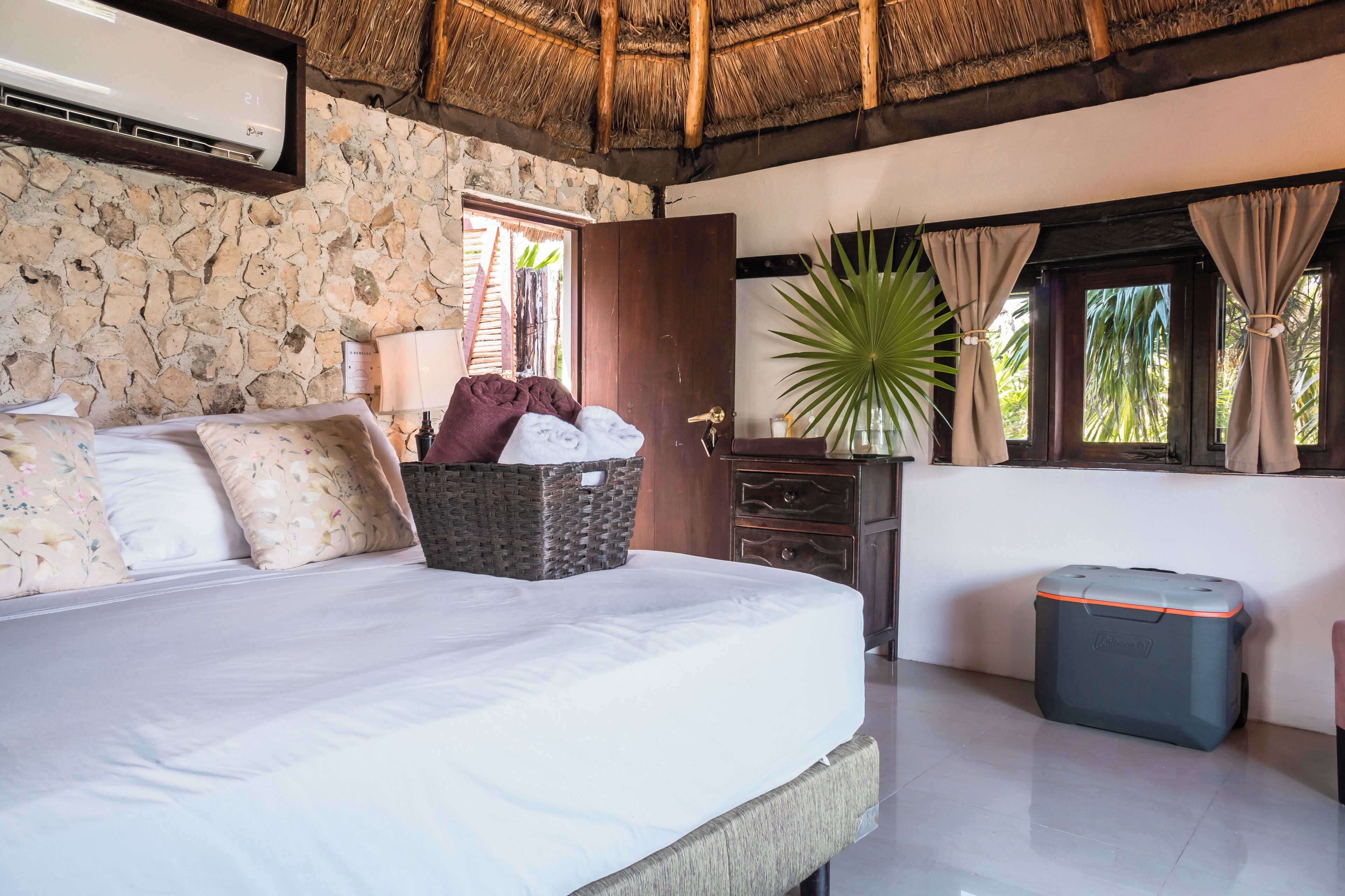 A bedroom with a stone wall, a bed with floral pillows, a wicker basket, a nightstand, a lamp, a potted plant, a window with curtains, and a suitcase.