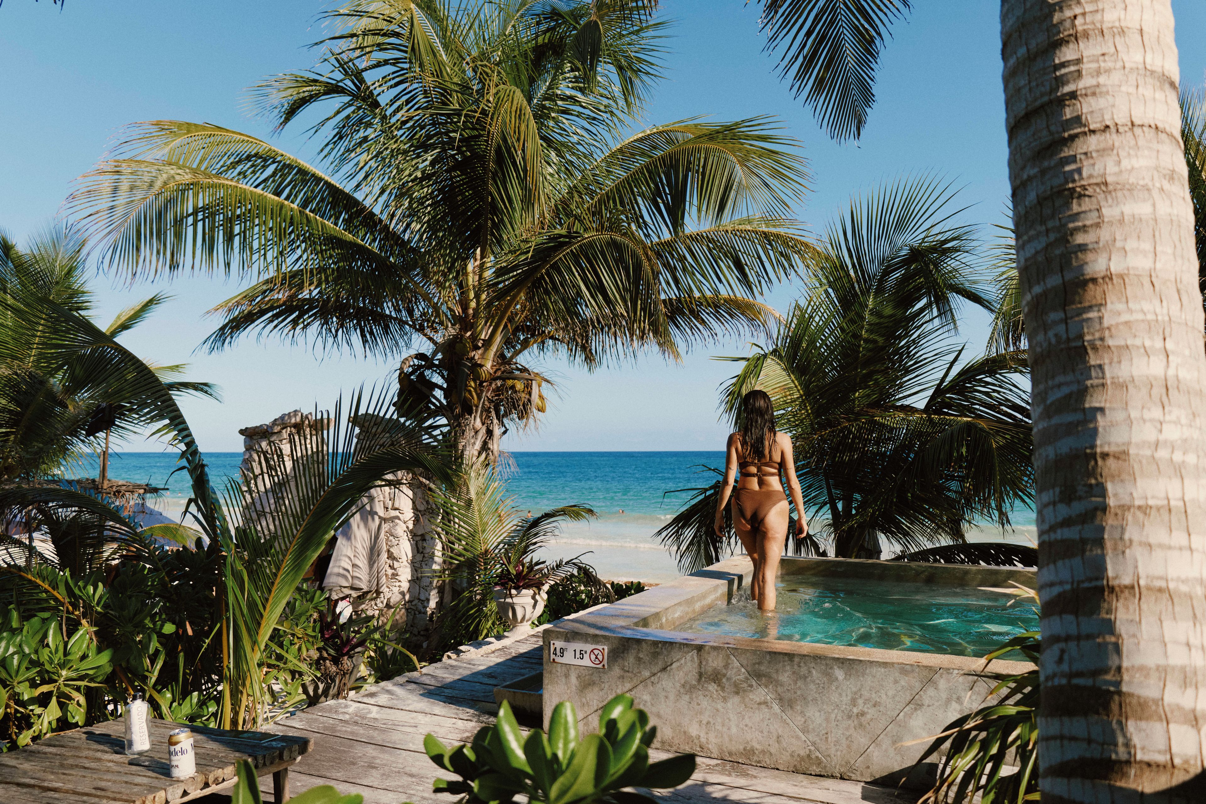 A pool area with a person standing by the edge, surrounded by palm trees and a view of the ocean.