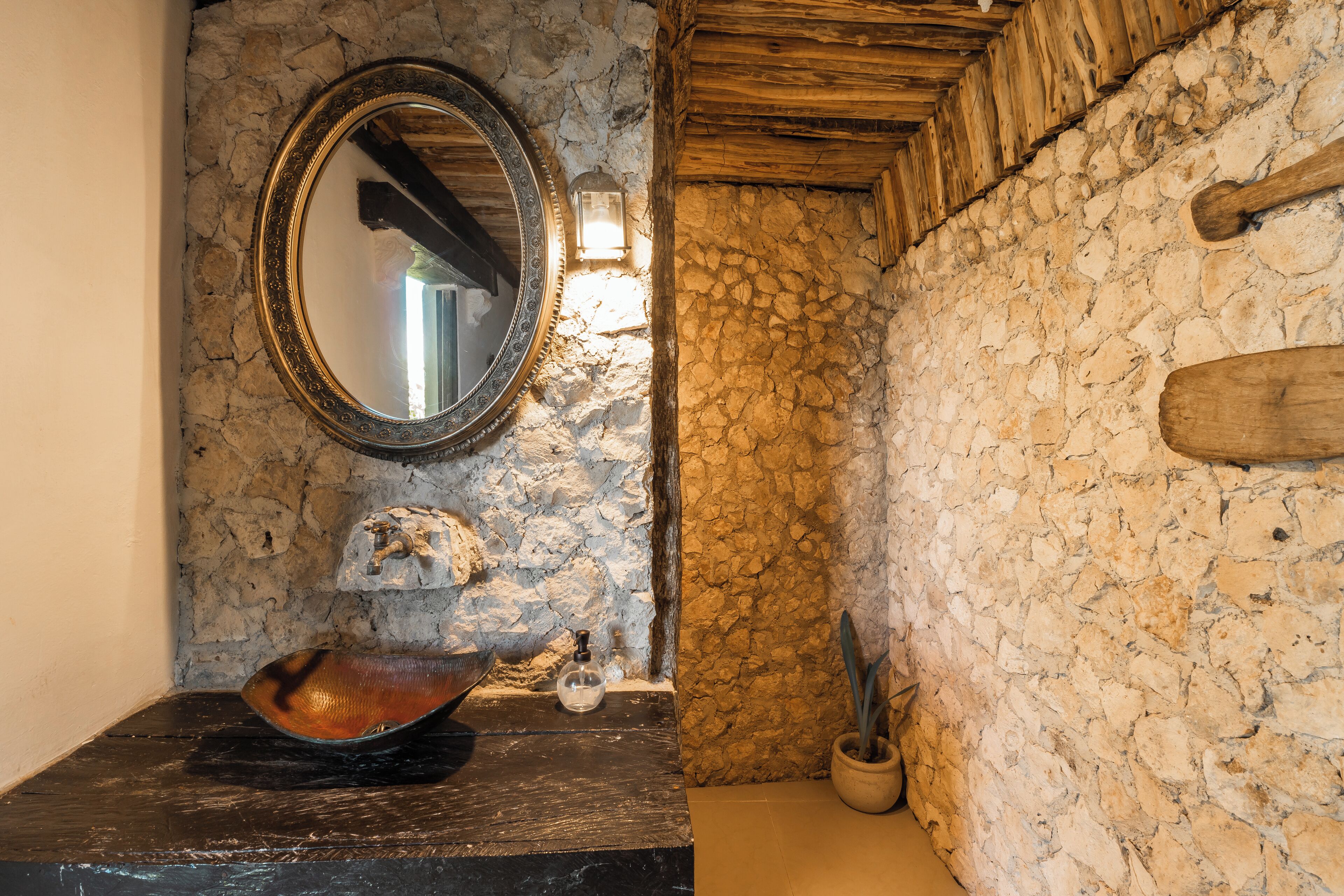 A rustic bathroom with a stone wall, a wooden ceiling, a round mirror, a black sink, a decorative bowl, a potted plant, and a wooden bathtowel rack.
