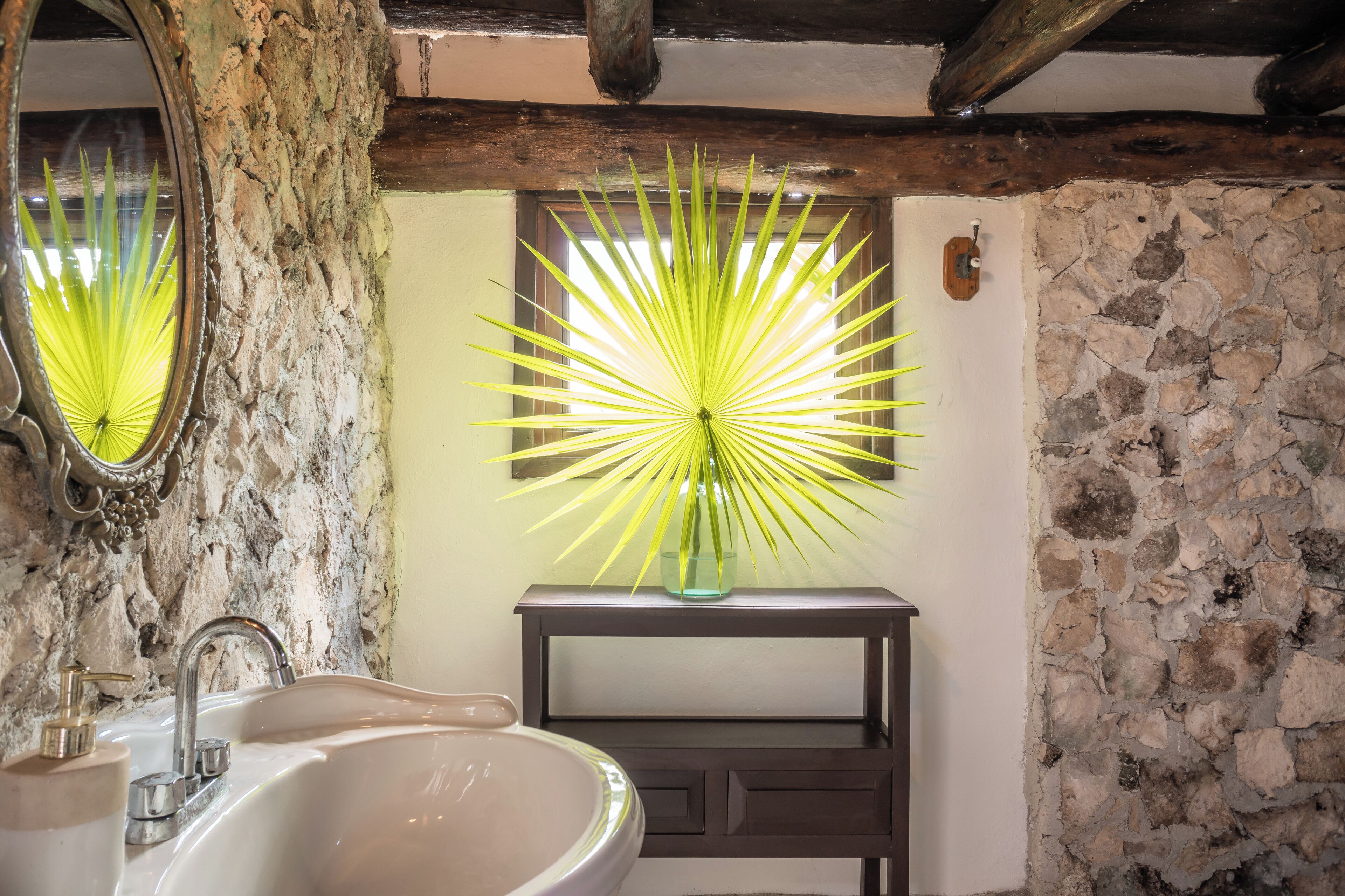 A bathroom with a stone wall, a wooden beam ceiling, a sink with a faucet, and a vase with a plant.