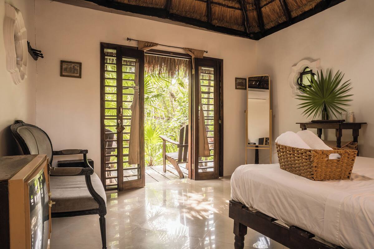 A bedroom with a bed, a chair, a wooden desk, a wicker basket, a mirror, and a plant.