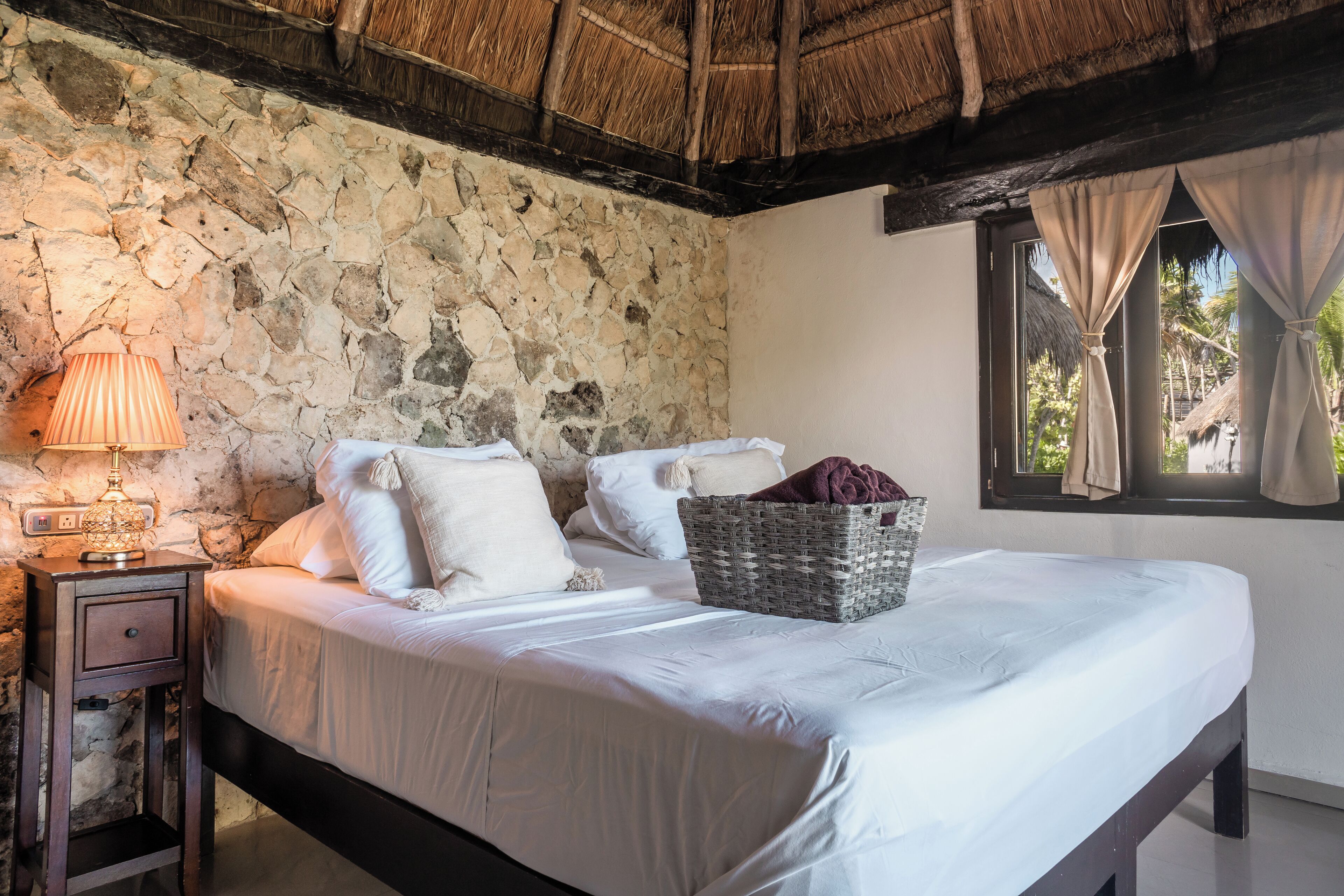 Une chambre avec un mur en pierre, un lit recouvert de draps blancs, une table de chevet avec une lampe et une fenêtre avec des rideaux.