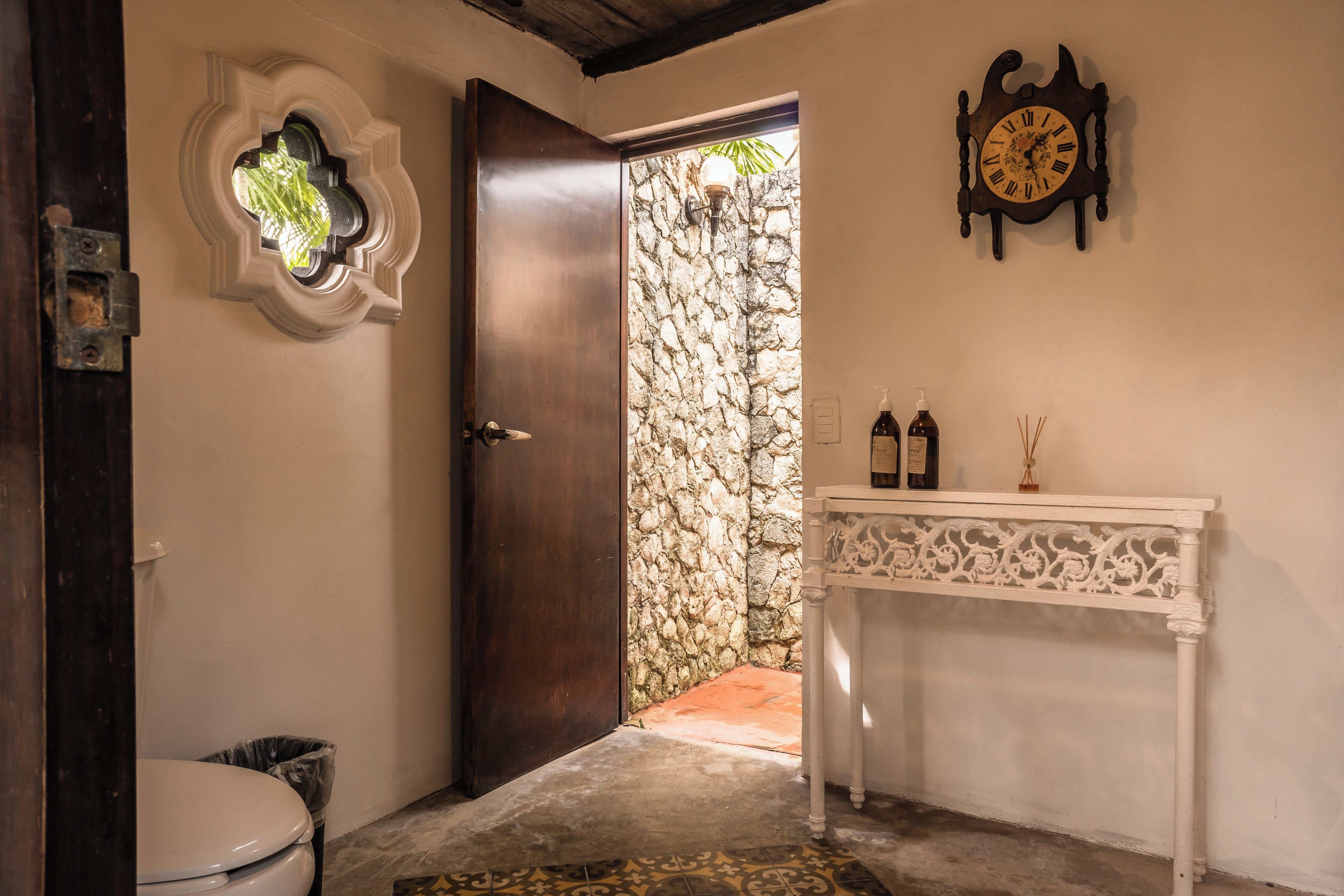 A bathroom with a stone wall, a wooden door, a white sink with bottles, and a wall clock.