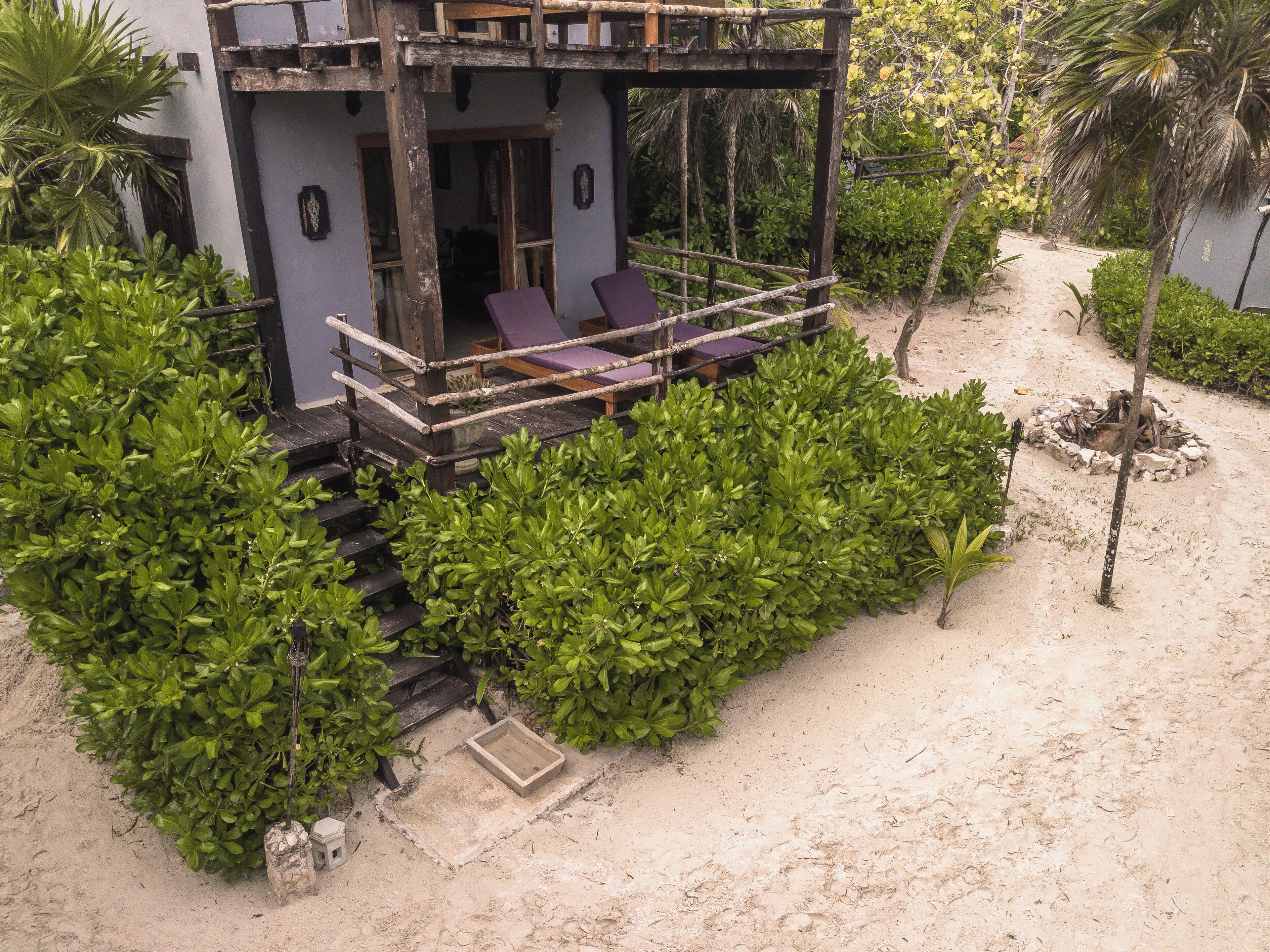 A two-story building with a balcony, surrounded by dense greenery and a sandy area.
