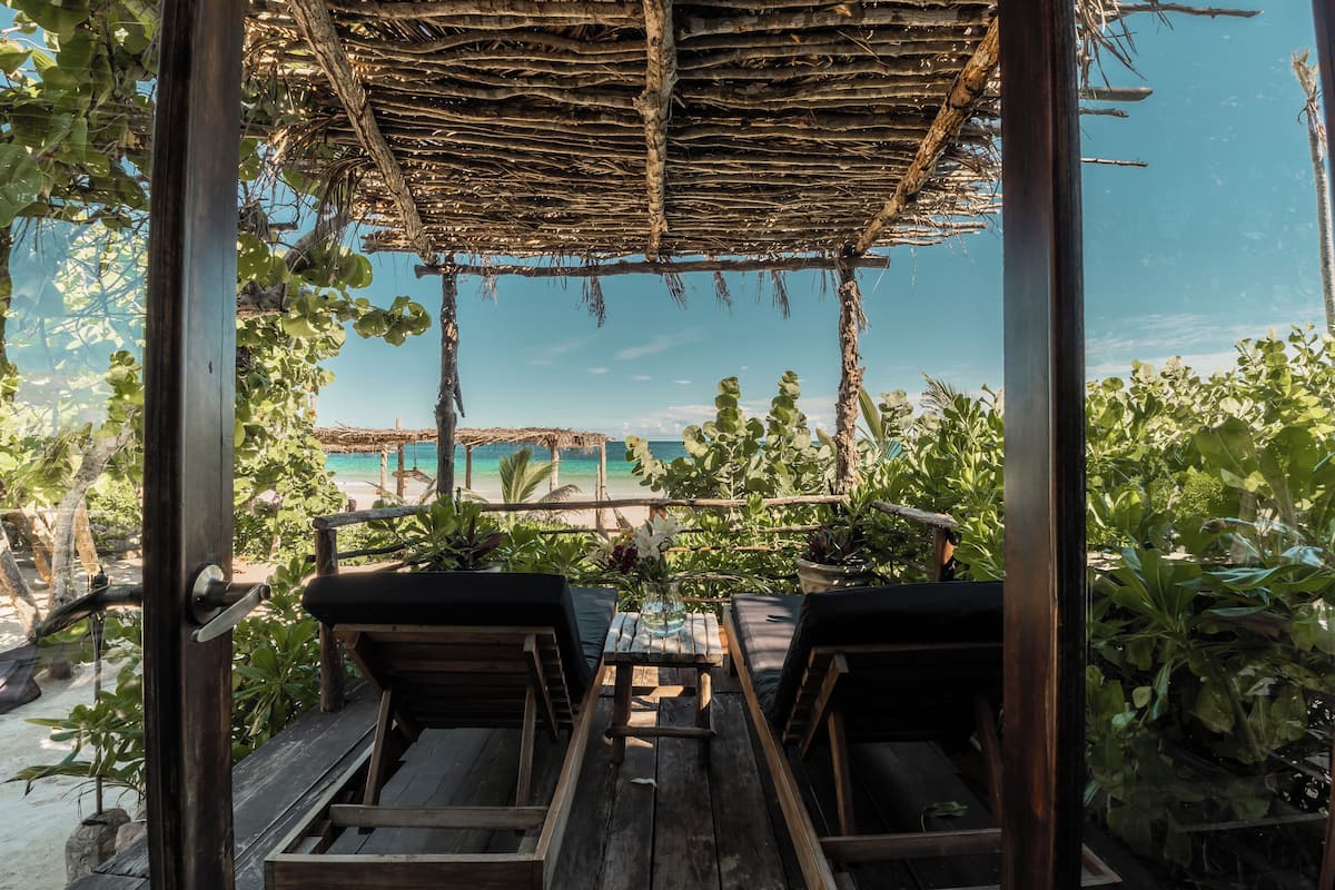 A wooden deck with two lounge chairs, a glass table, and a thatched roof structure overlooking a beach.