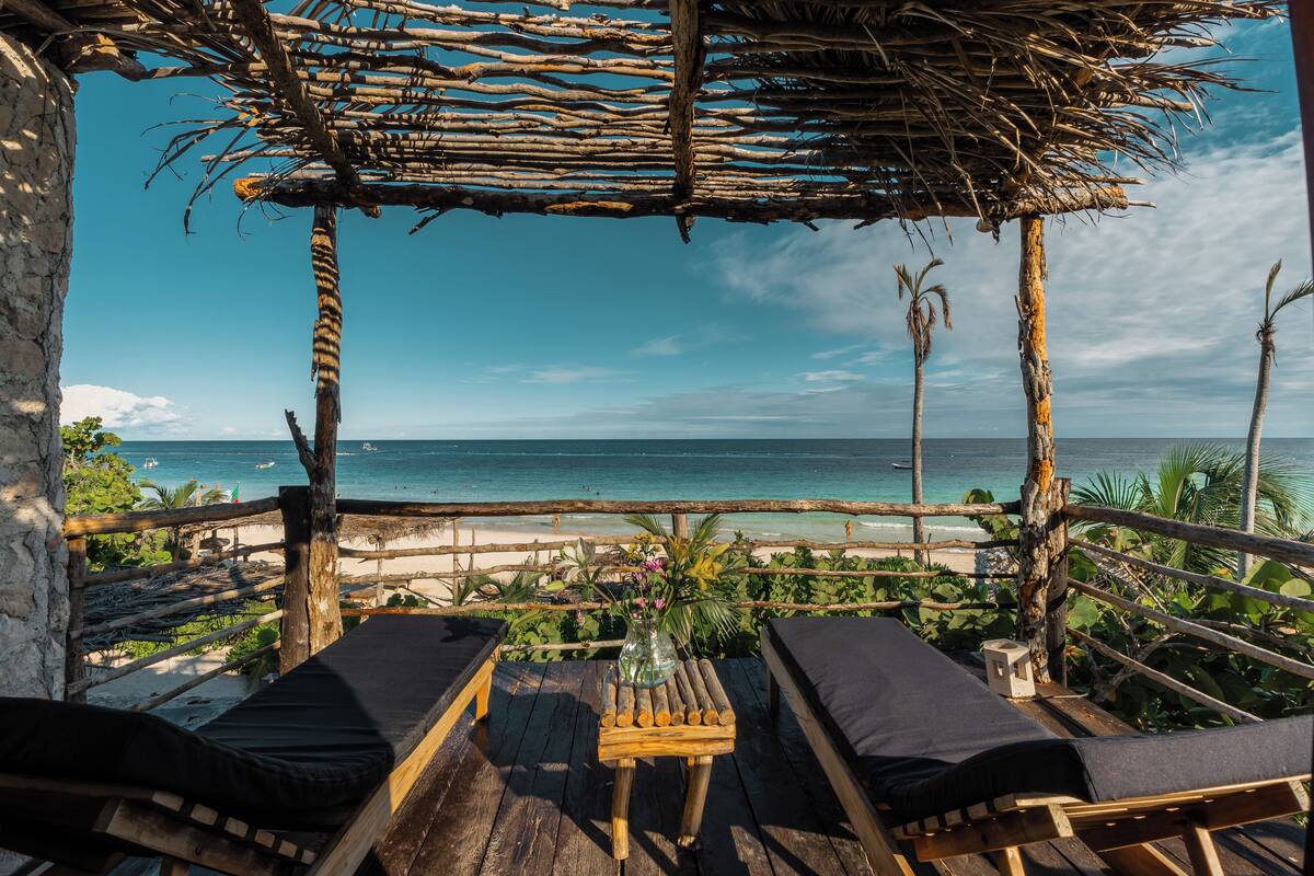A wooden deck with two lounge chairs and a table, overlooking a beach with palm trees and the ocean.