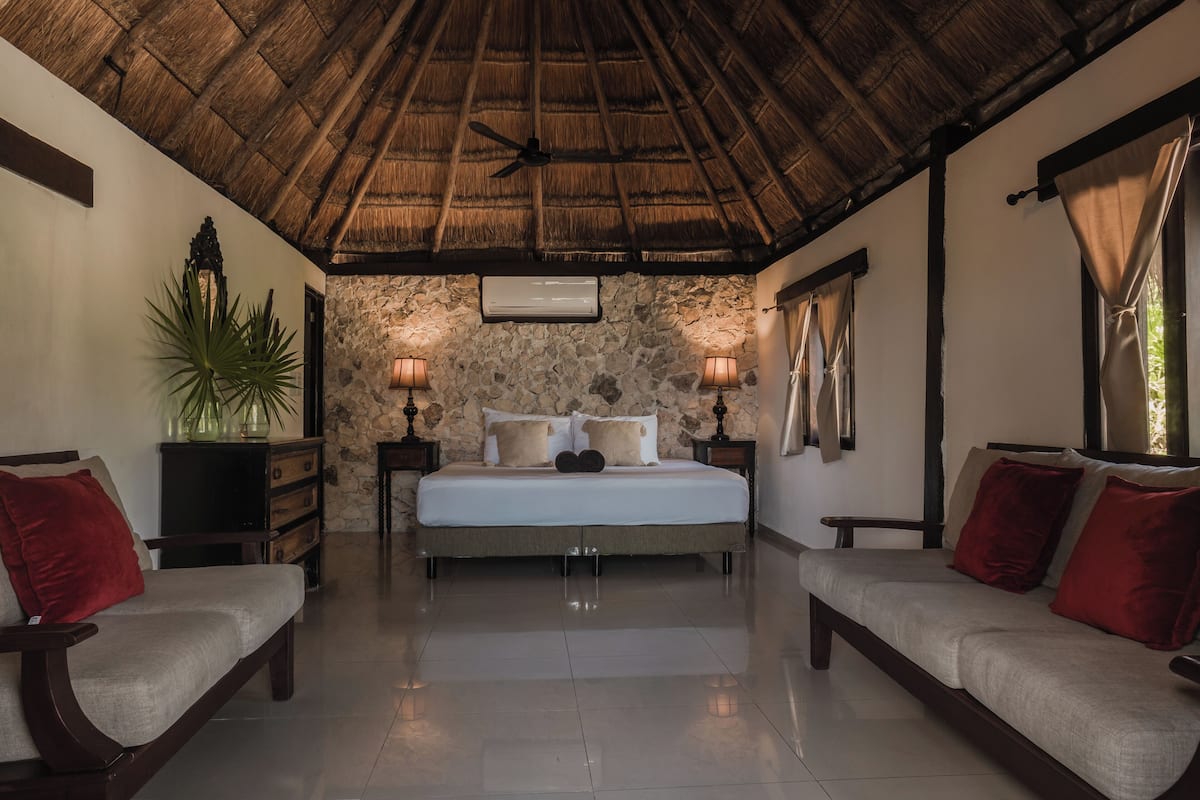 A bedroom with a stone wall, a bed with two pillows, a nightstand with a lamp, a ceiling fan, and a potted plant.