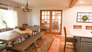 Comedor con mesa de madera, bancos y una araña. Se ve un salón a través de una puerta de vidrio.