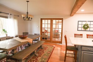 Zona pranzo con tavolo in legno, panche e lampadario. Si vede un soggiorno attraverso una porta in vetro.