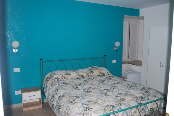 A bedroom with a teal wall, a bed with floral bedding, a nightstand, a small bedside table, and a wardrobe.
