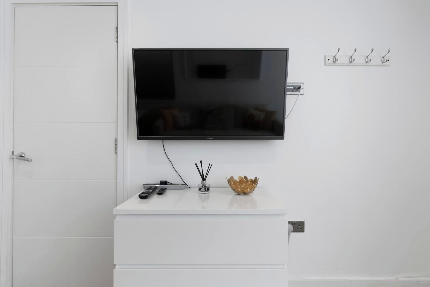 Um armário branco com uma televisão montada acima, um controle remoto e uma tigela decorativa no balcão.