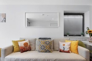 A living room with a sofa, cushions, a mirror, and a window with a view.
