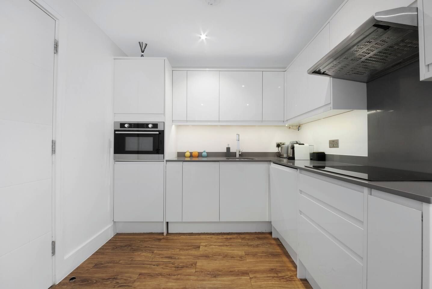 Una cucina moderna con mobili bianchi, forno integrato, cappa aspirante in acciaio inossidabile e pavimento in legno.