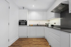 A modern kitchen with white cabinetry, a built-in oven, a stainless steel range hood, and a wooden floor.