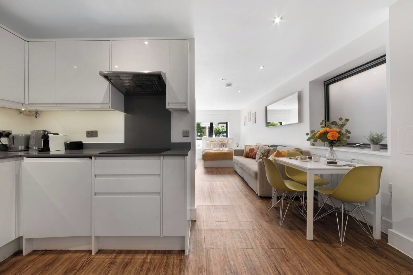 A modern kitchen with white cabinetry, a black range hood, and a dining area with a white table and yellow chairs.
