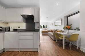 A modern kitchen with white cabinetry, a black range hood, and a dining area with a white table and yellow chairs. - Hermitage Rd N4, Manor House | Ground floor (London)