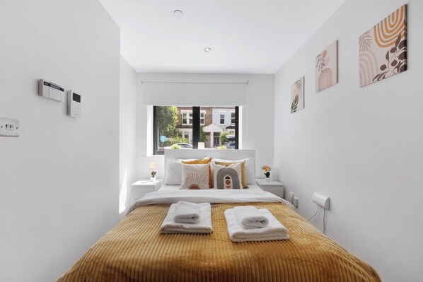 Une chambre d’hôtel moderne avec un grand lit, une fenêtre donnant sur des immeubles, une table de chevet avec une lampe et un climatiseur fixé au mur.