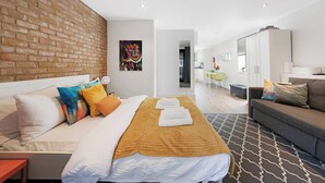 Ein modernes Hotelzimmer mit Ziegelwand, einem Bett mit vielen Kissen, einer Couch, einem Essbereich und einer Küche im Hintergrund.