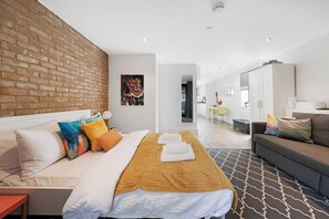 Quarto de hotel moderno com parede de tijolos decorativa, cama com várias almofadas, sofá, área de refeições e cozinha ao fundo.