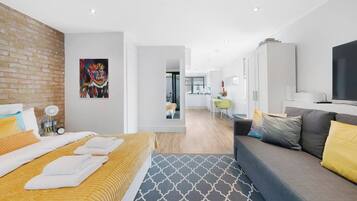 A modern hotel room with a bed, a sofa, a TV, and a kitchen area in the background.