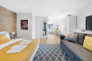 A modern hotel room with a bed, a sofa, a TV, and a kitchen area in the background.