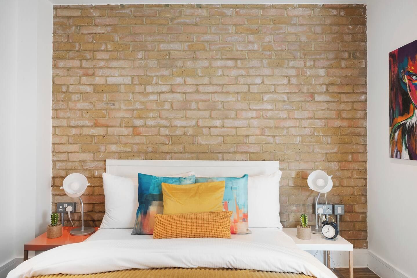 A bedroom with a brick wall, a white bed, colorful pillows, bedside tables with lamps, and a cactus plant.