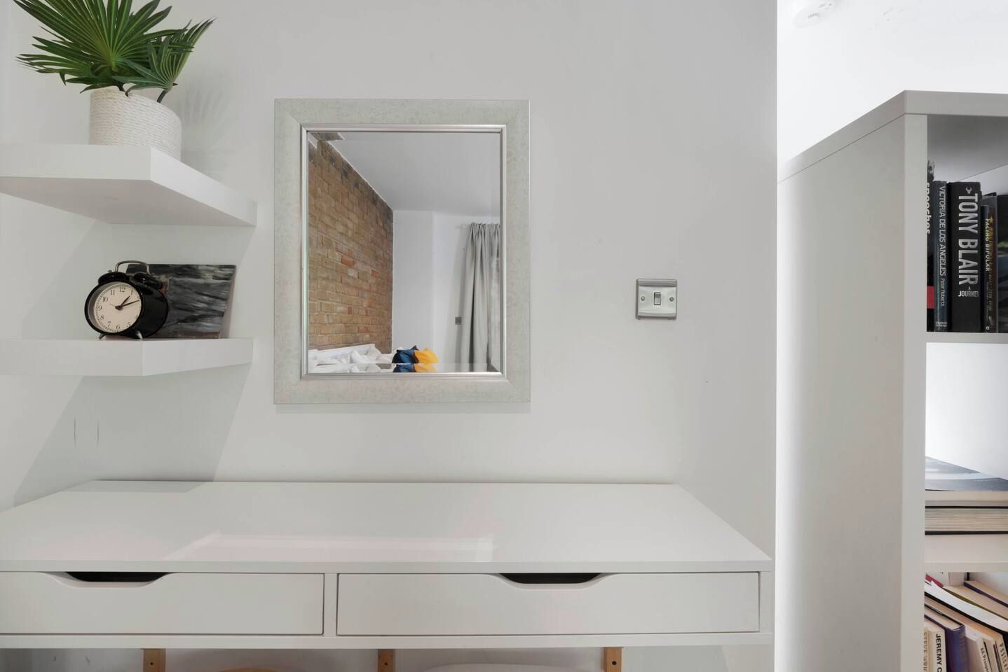 A white desk with drawers, a mirror, a potted plant, a clock, and a bookshelf with books.