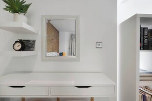 Un bureau blanc avec des tiroirs, un miroir, une plante en pot, une horloge et une bibliothèque remplie de livres.