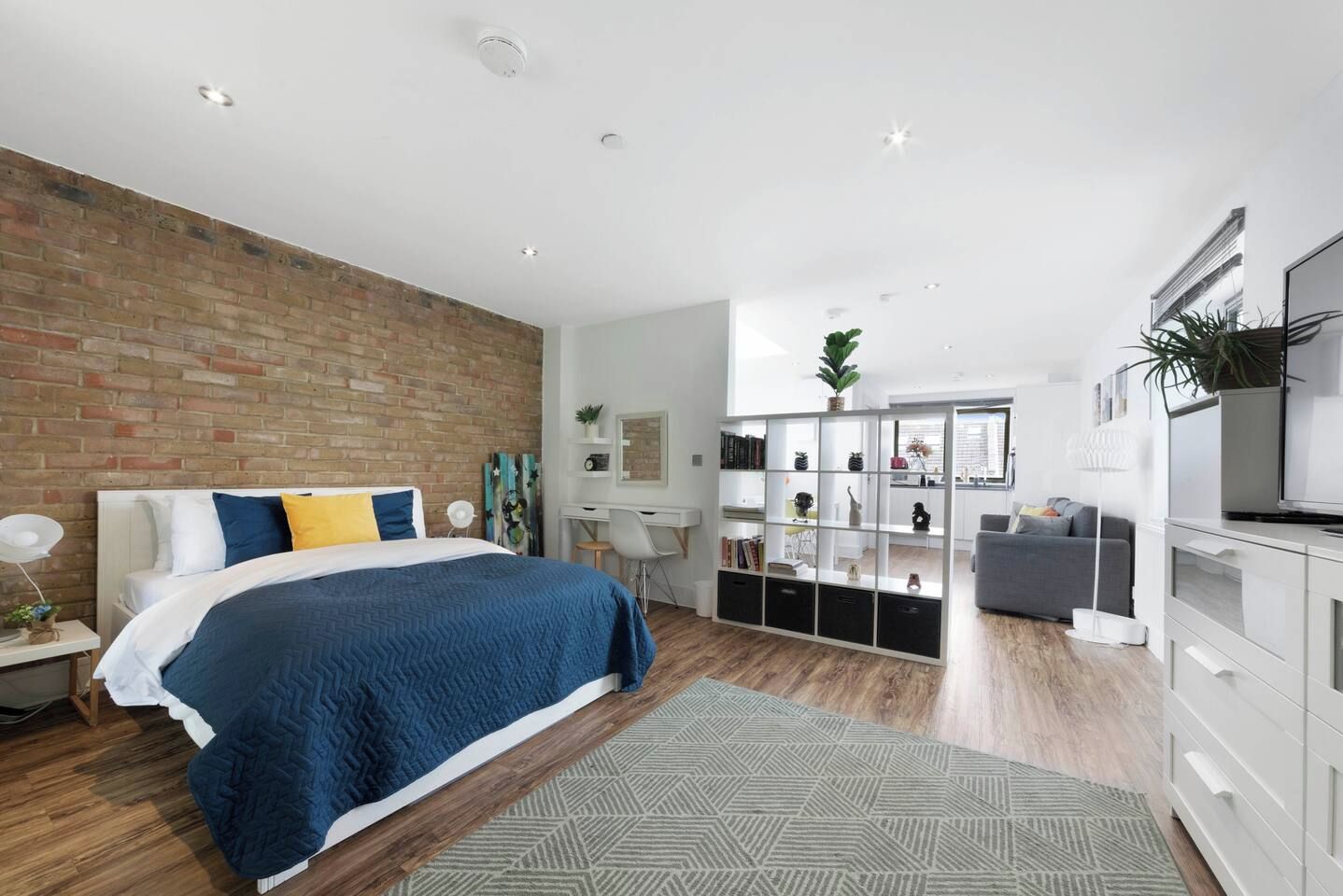 A modern bedroom with a brick accent wall, a large bed with blue bedding, a desk, and a grey sofa.