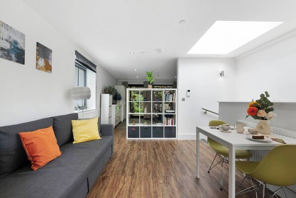 A modern living room with a grey sofa, a dining table with chairs, and a bookshelf.