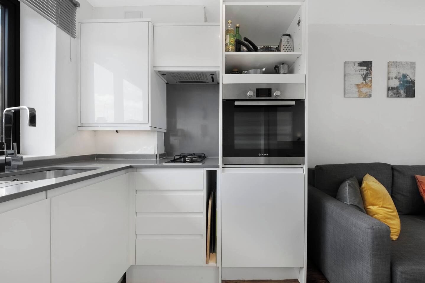 Eine kompakte Küche mit weißen Schränken, einem Bosch-Ofen und einem grauen Sofa mit gelben Kissen.
